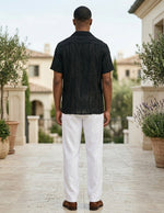 Man wearing a black shirt and white pants standing in an outdoor setting with plants and buildings in the background.