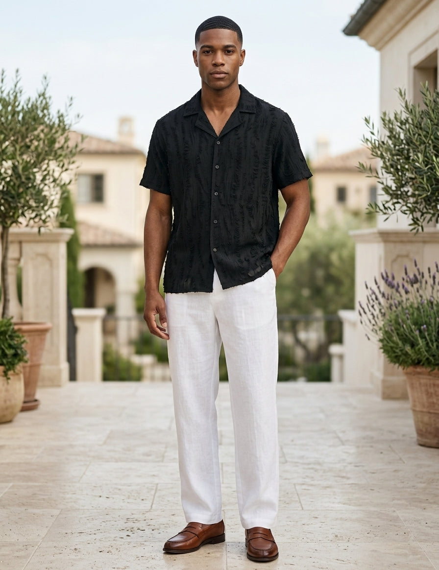 Man wearing a black shirt and white pants standing outdoors with plants and buildings in the background