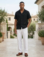 Man wearing a black shirt and white pants standing outdoors with plants and buildings in the background