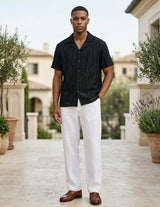 Man wearing a black shirt and white pants standing outdoors with plants and buildings in the background