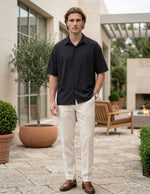 Man wearing a black shirt and white pants standing outdoors in a patio setting.