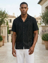 Man wearing a black short-sleeve shirt and white pants standing outdoors.