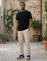 Man wearing a black t-shirt and beige pants standing in an outdoor setting with stone walls and plants.