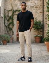 Man wearing a black t-shirt and beige pants standing in an outdoor setting with stone walls and plants.