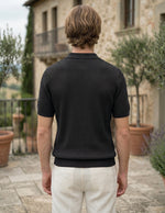 Man wearing a black textured polo shirt and white pants standing in front of a stone building with greenery.