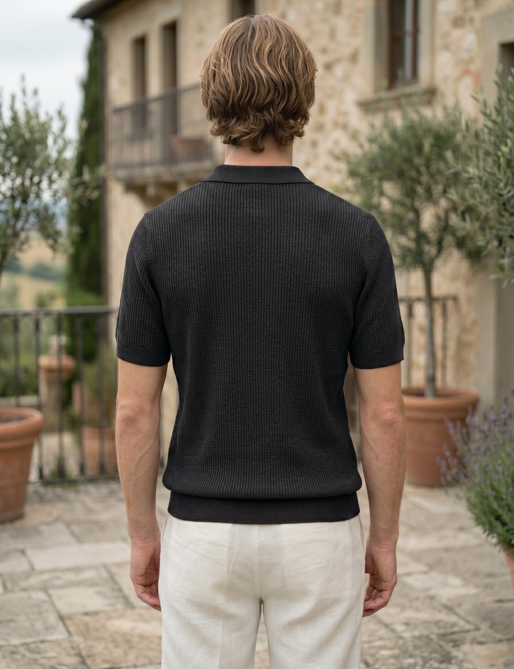 Man wearing a black textured polo shirt and white pants standing in front of a stone building with greenery.