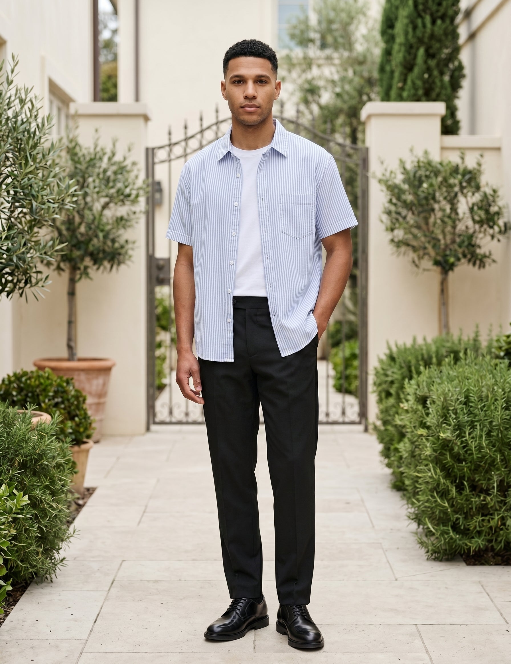 Man wearing a blue-and-white striped short-sleeve shirt over a white T-shirt with black trousers and black shoes outdoors
