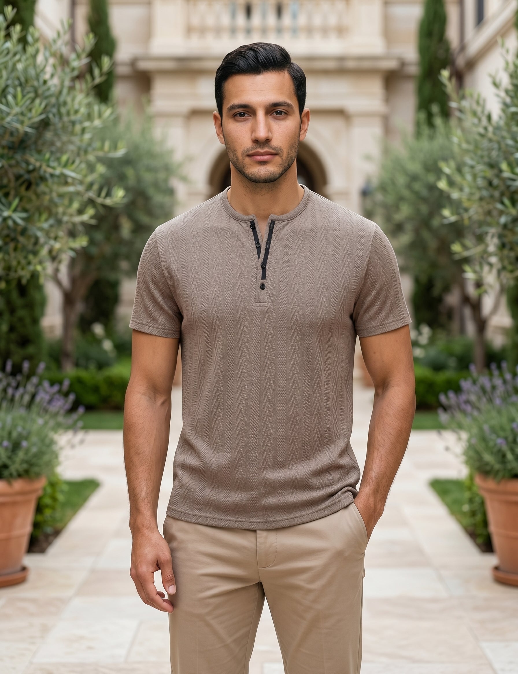 Man wearing a brown henley shirt and beige pants standing in a garden setting.