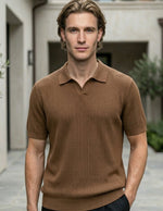 Man wearing a brown polo shirt standing in an outdoor setting with plants and a building.