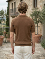 Man wearing a brown textured shirt and white pants standing in an outdoor setting with stone building and potted plants.