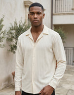 Man wearing a cream-colored button-up shirt standing outdoors with plants and a building in the background.