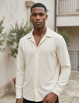 Man wearing a cream-colored button-up shirt standing outdoors with plants and a building in the background.