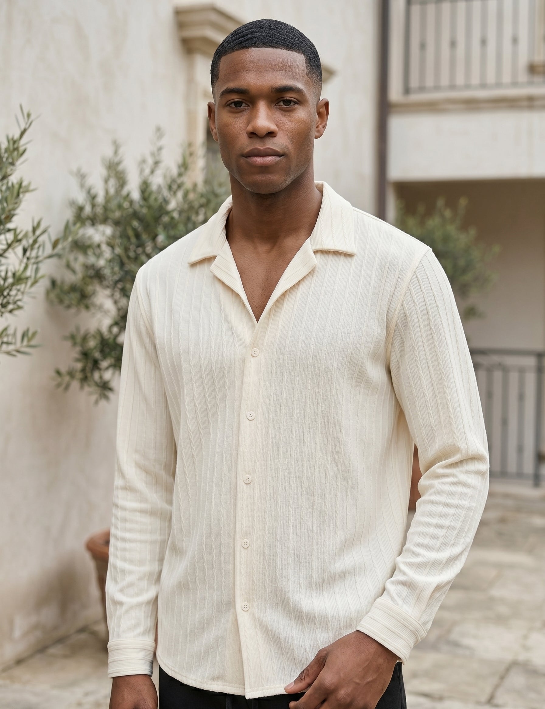 Man wearing a cream-colored button-up shirt standing outdoors with plants and a building in the background.