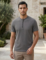 Man wearing a gray short-sleeve shirt standing outdoors with a stone building and plants in the background