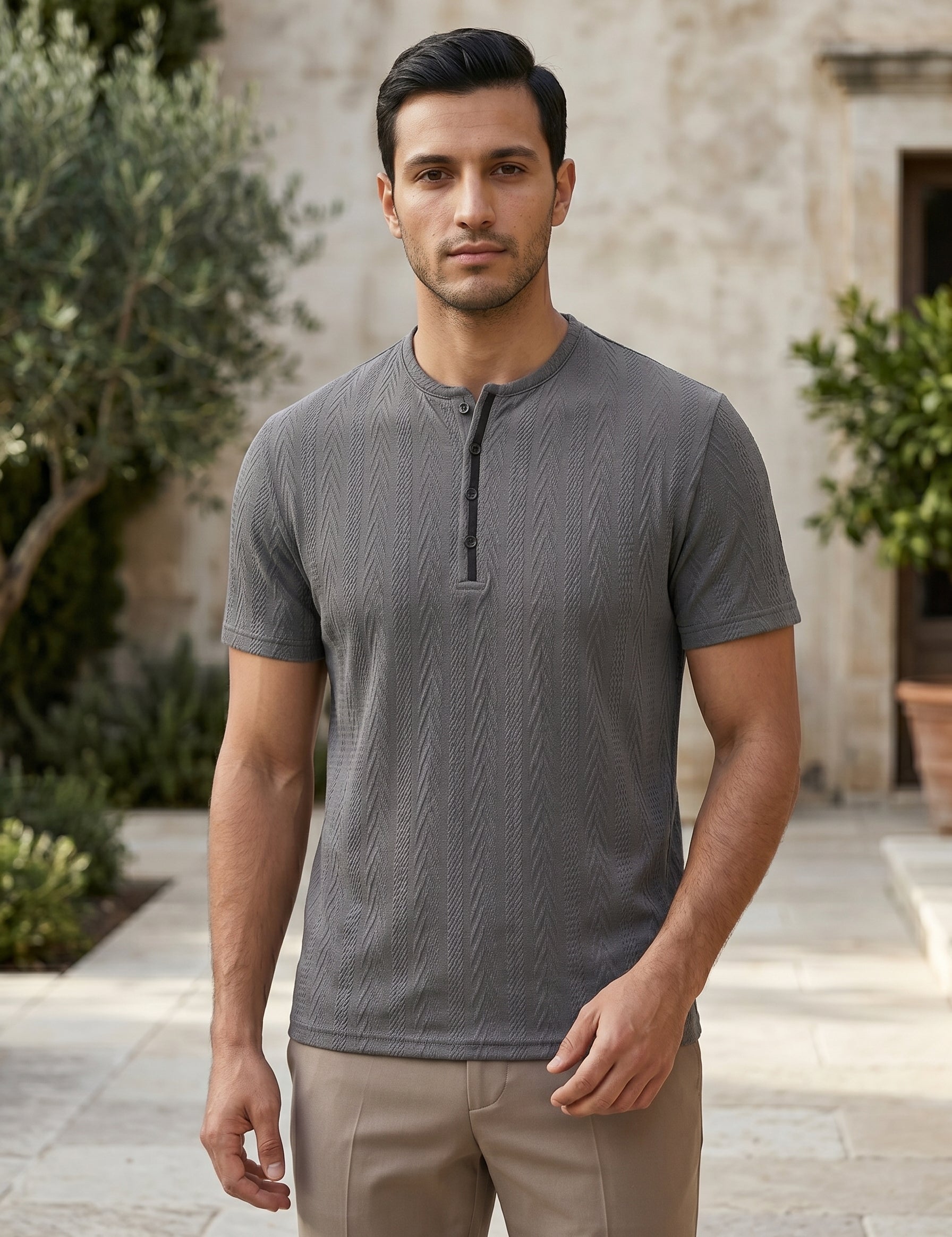 Man wearing a gray short-sleeve shirt standing outdoors with a stone building and plants in the background