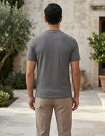 Man wearing a gray t-shirt and beige pants standing in an outdoor setting with plants and a building.