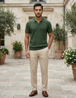 Man wearing a green polo shirt and beige pants standing in an outdoor setting with plants and a building.