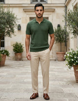 Man wearing a green polo shirt and beige pants standing in an outdoor setting with plants and a building.
