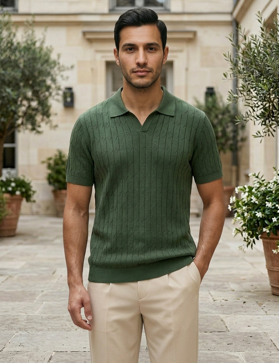 Man wearing a green polo shirt and beige pants standing in an outdoor setting with plants and a building.