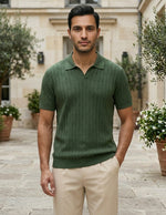 Man wearing a green polo shirt and beige pants standing in an outdoor setting with plants and a building.