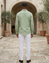 Man wearing a green shirt and white pants standing in an outdoor setting with potted plants and stone pavement.