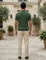 Man wearing a green sweater and beige pants standing in an outdoor setting with potted plants.