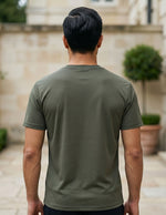 Man wearing a green t-shirt standing in an outdoor setting with a stone wall and plants.