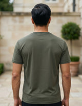 Man wearing a green t-shirt standing in an outdoor setting with a stone wall and plants.
