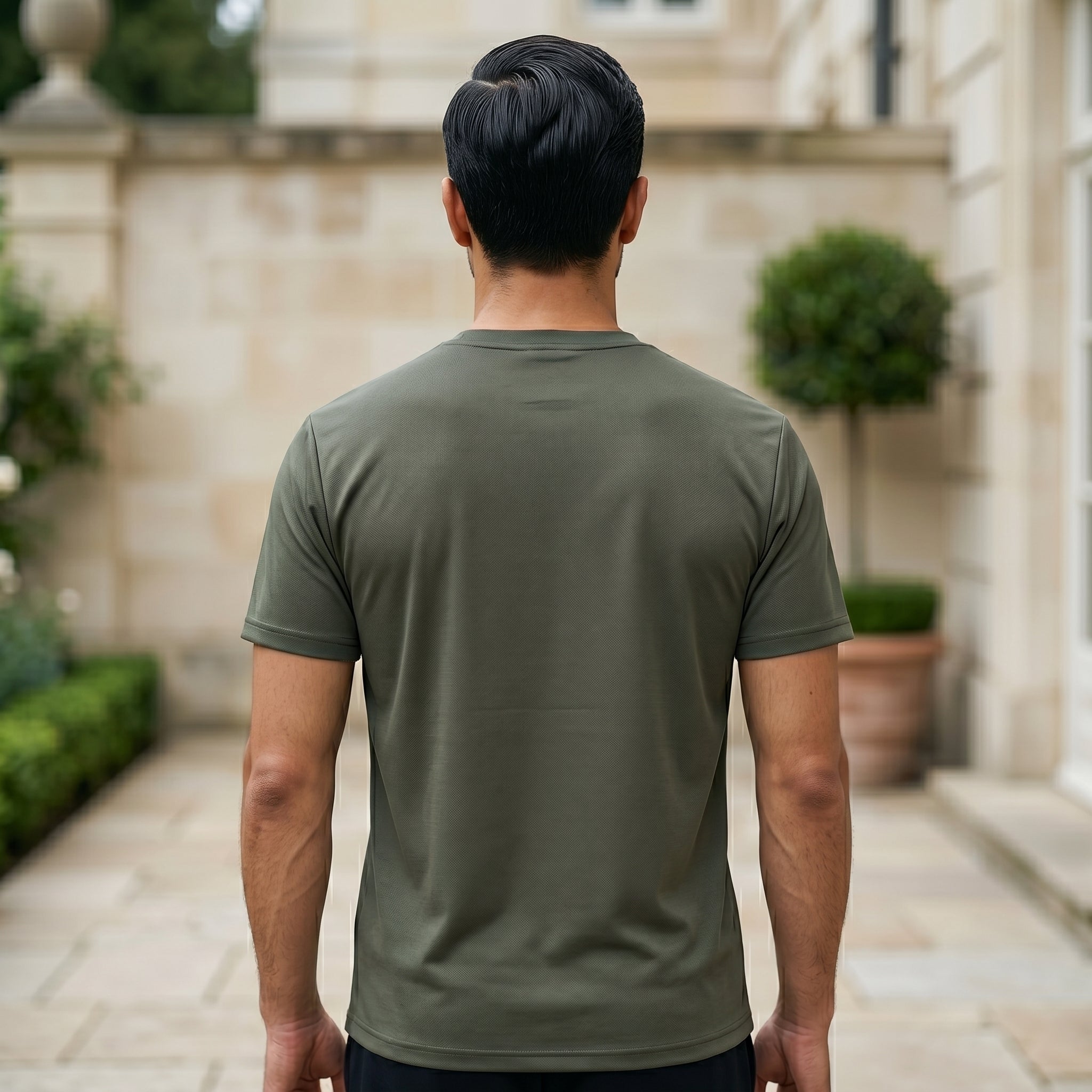 Man wearing a green t-shirt standing in an outdoor setting with a stone wall and plants.