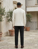 Man wearing a light-colored shirt and dark pants standing in an outdoor setting with plants and a building.