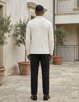Man wearing a light-colored shirt and dark pants standing in an outdoor setting with plants and a building.