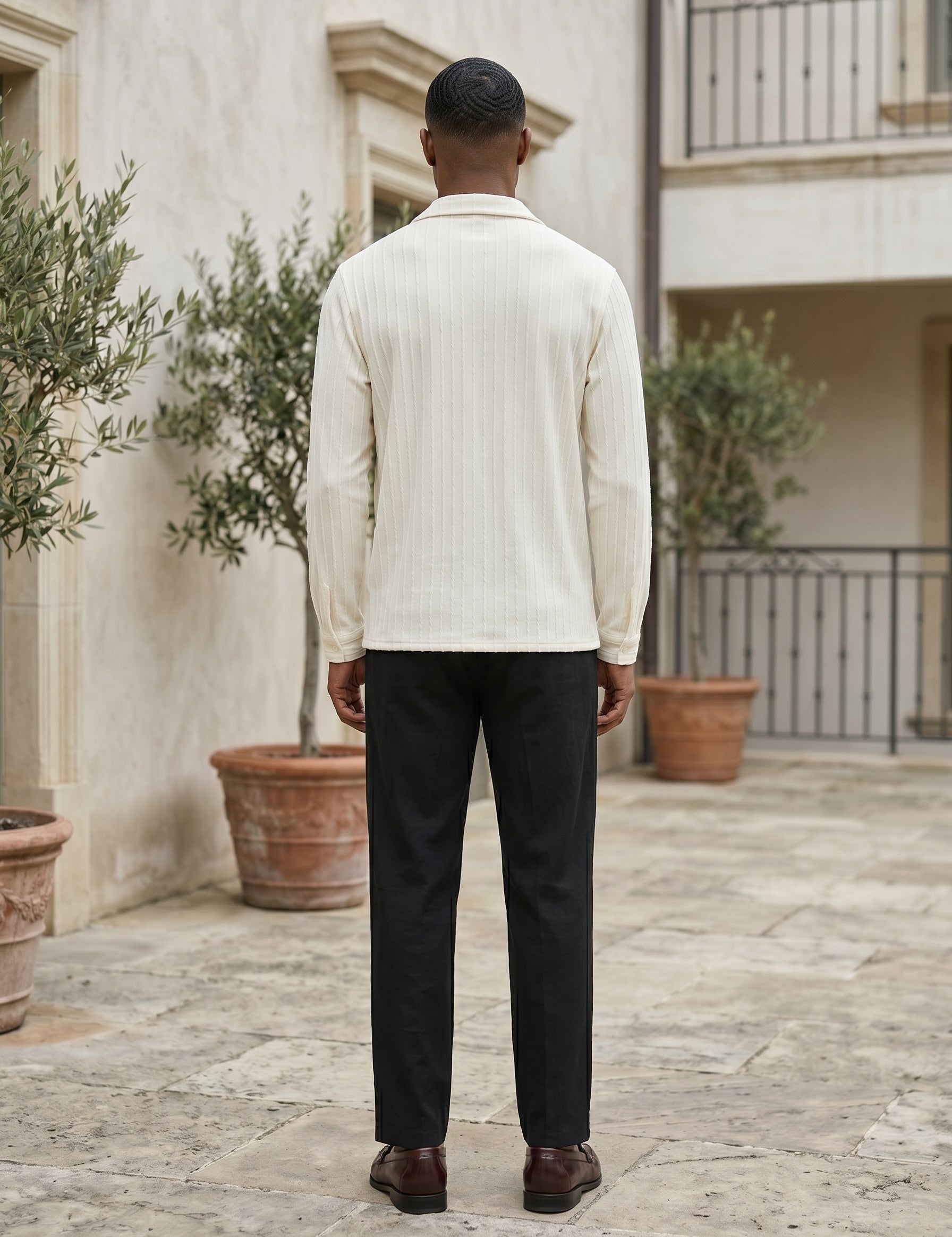 Man wearing a light-colored shirt and dark pants standing in an outdoor setting with plants and a building.