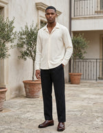 Man wearing a light-colored shirt and dark pants standing in an outdoor setting with plants and a building in the background.