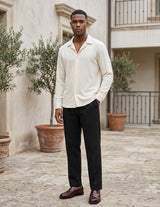 Man wearing a light-colored shirt and dark pants standing in an outdoor setting with plants and a building in the background.