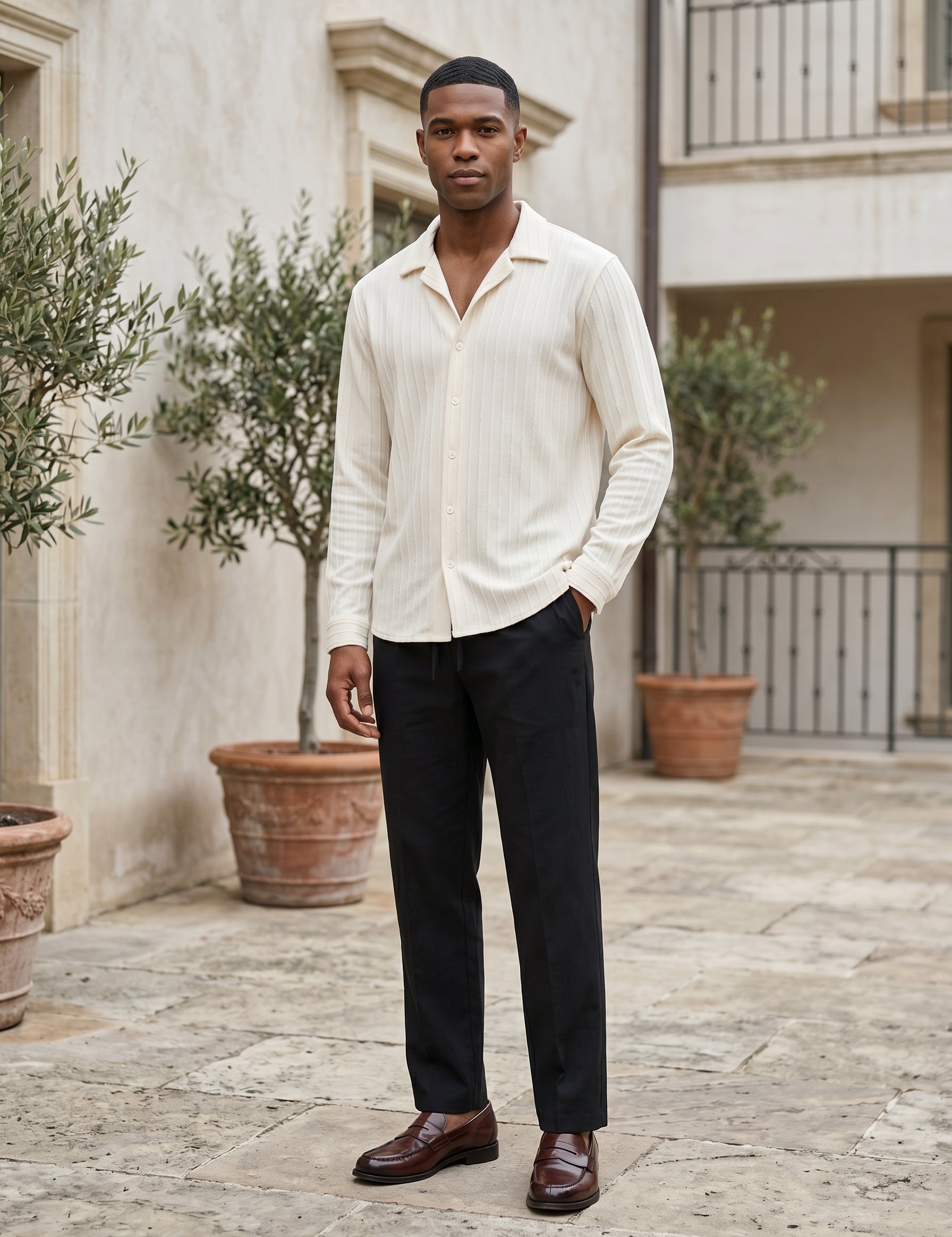 Man wearing a light-colored shirt and dark pants standing in an outdoor setting with plants and a building in the background.