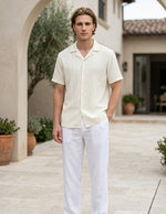 Man wearing a light-colored shirt and white pants standing outdoors.