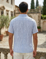 Man wearing a light blue patterned shirt standing in an outdoor setting with buildings and trees.