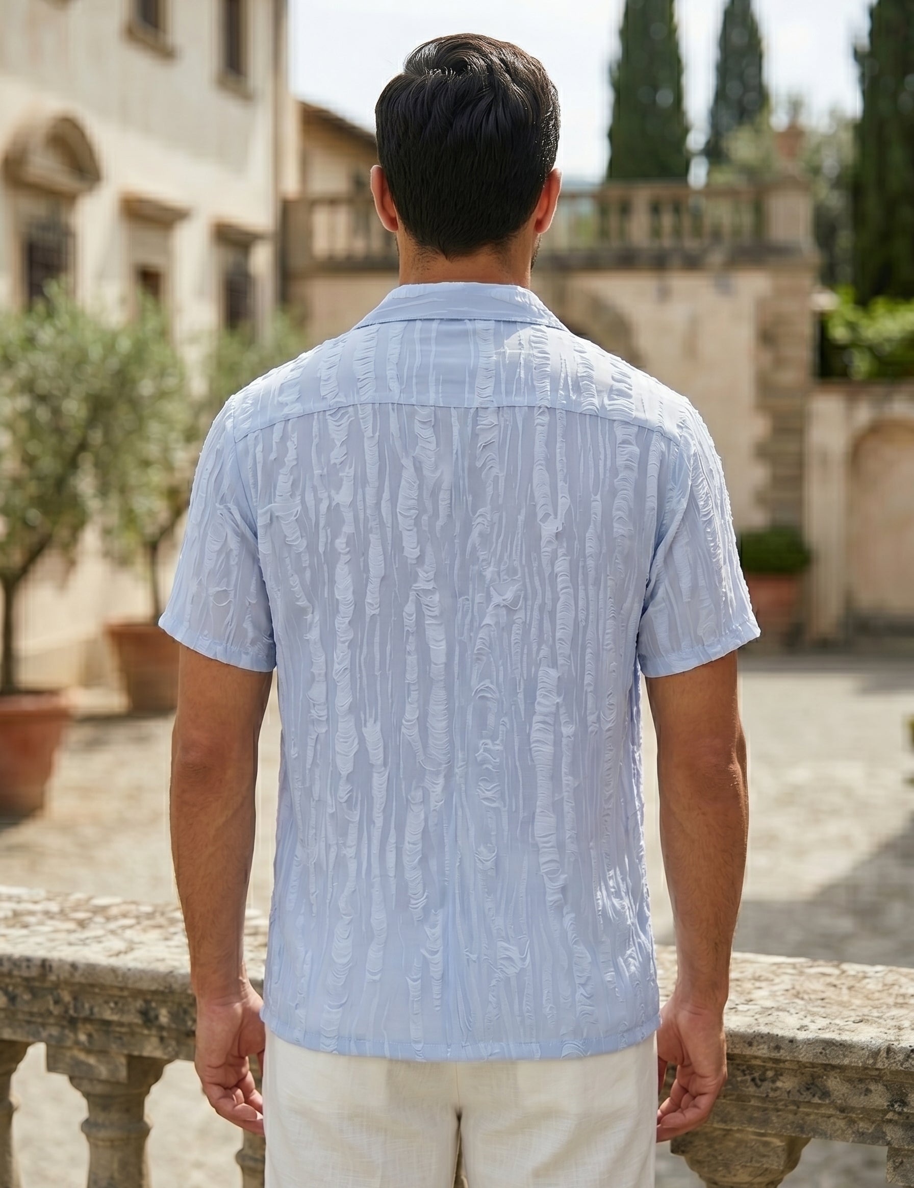 Man wearing a light blue patterned shirt standing in an outdoor setting with buildings and trees.
