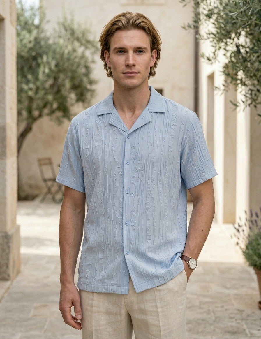 Man wearing a light blue patterned shirt standing outdoors with a building and trees in the background