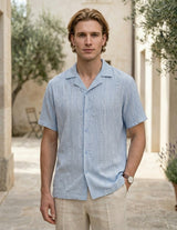 Man wearing a light blue patterned shirt standing outdoors with a building and trees in the background