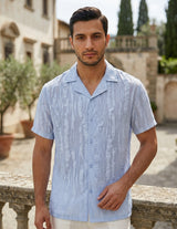 Man wearing a light blue patterned shirt standing outdoors with a stone wall and greenery in the background.