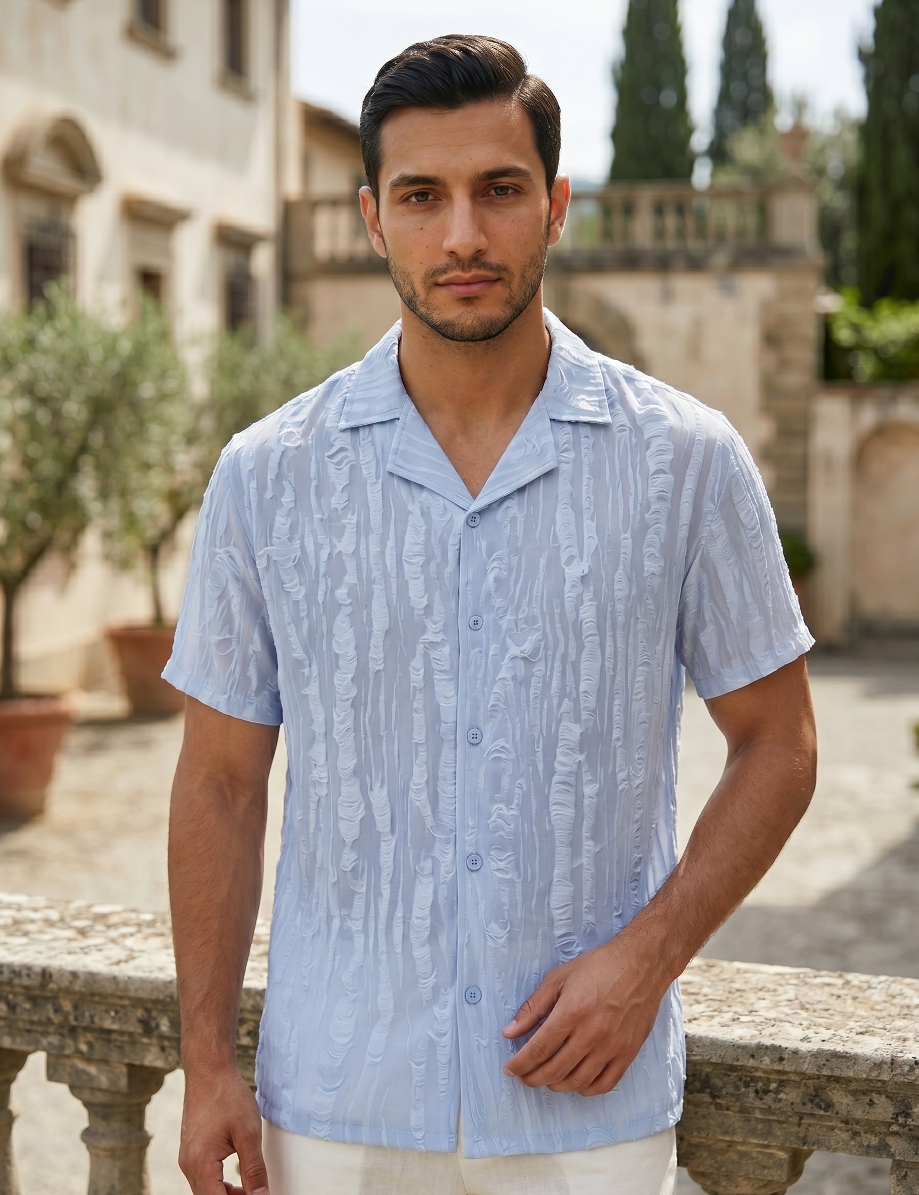 Man wearing a light blue patterned shirt standing outdoors with a stone wall and greenery in the background.