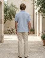 Man wearing a light blue shirt and beige pants standing in an outdoor setting with stone walls and potted plants.