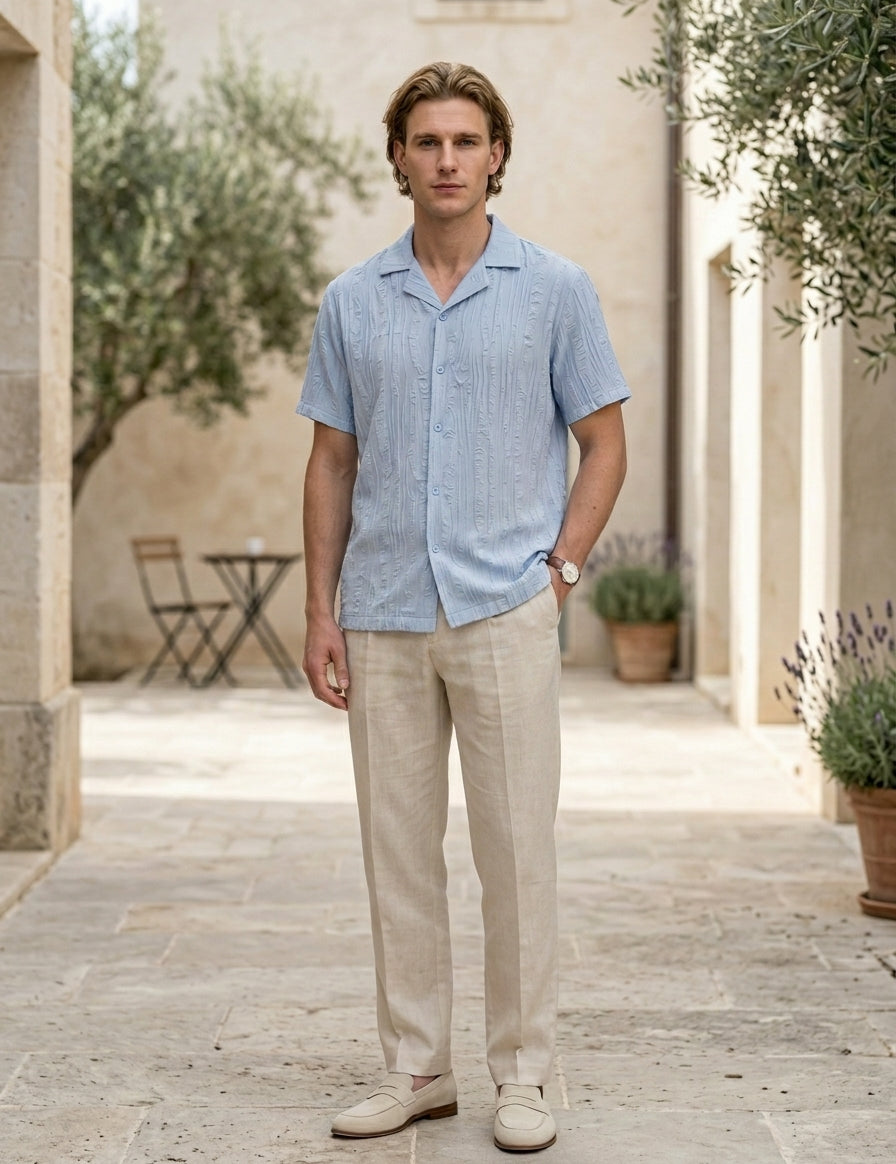 Man wearing a light blue shirt and beige pants standing in an outdoor setting with plants and a table.