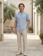 Man wearing a light blue shirt and beige pants standing in an outdoor setting with plants and a table.