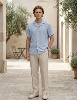 Man wearing a light blue shirt and beige pants standing in an outdoor setting with plants and a table.