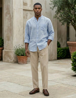 Man wearing a light blue shirt and beige pants standing in an outdoor setting with plants and stone pavement.