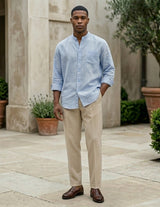 Man wearing a light blue shirt and beige pants standing in an outdoor setting with plants and stone pavement.