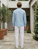 Man wearing a light blue shirt and white pants standing in an outdoor setting with plants and a building.