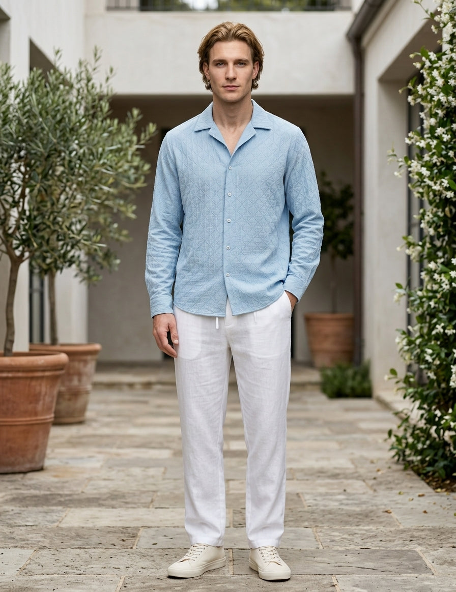 Man wearing a light blue shirt and white pants standing in an outdoor setting with plants and a building.
